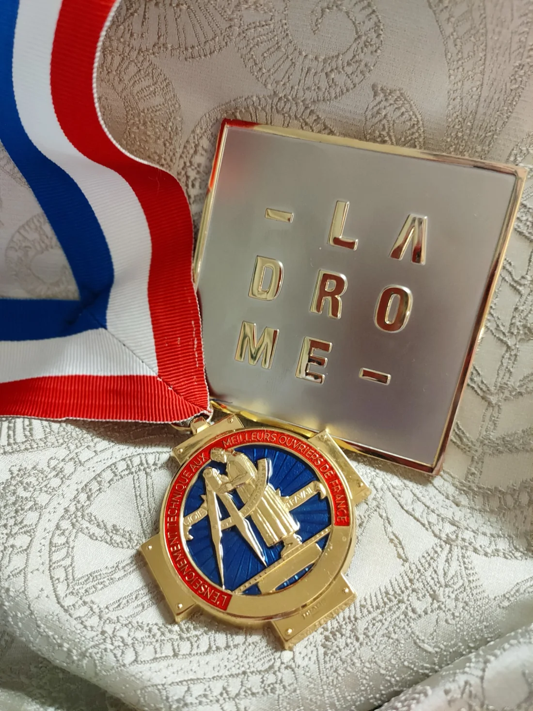 Médaille des Meilleurs Ouvriers de France et son ruban tricolore, posée sur un tissu damassé.