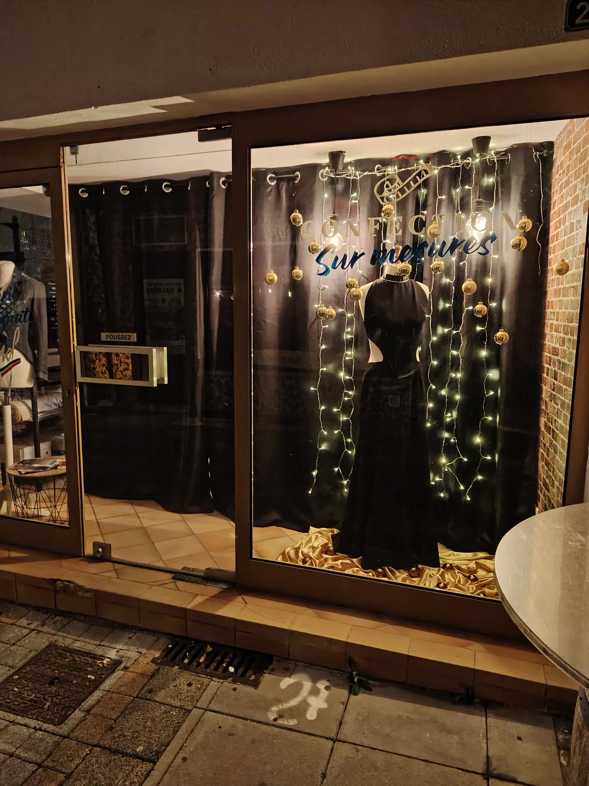 Vitrine de l'atelier éclairée la nuit, robe noire longue et guirlandes lumineuses.