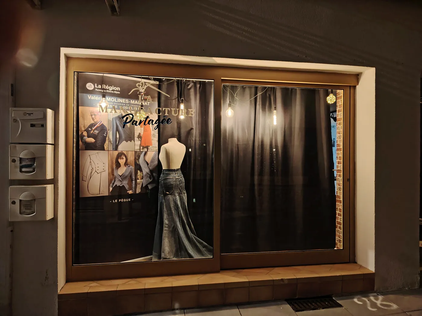 Façade de l'atelier la nuit, vitrine éclairée avec une robe noire et des tissus dorés.
